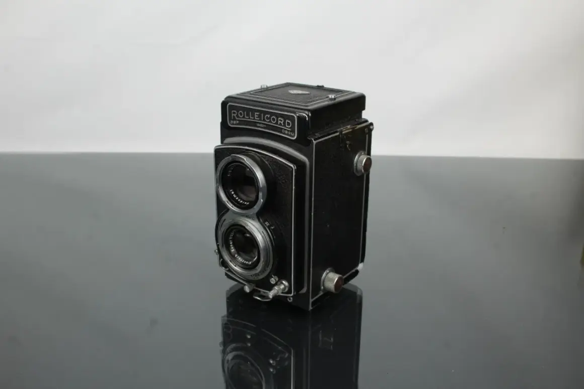 A vintage Rolleicord camera sits on a reflective surface, its twin lenses and classic design hinting at a rich photographic history.