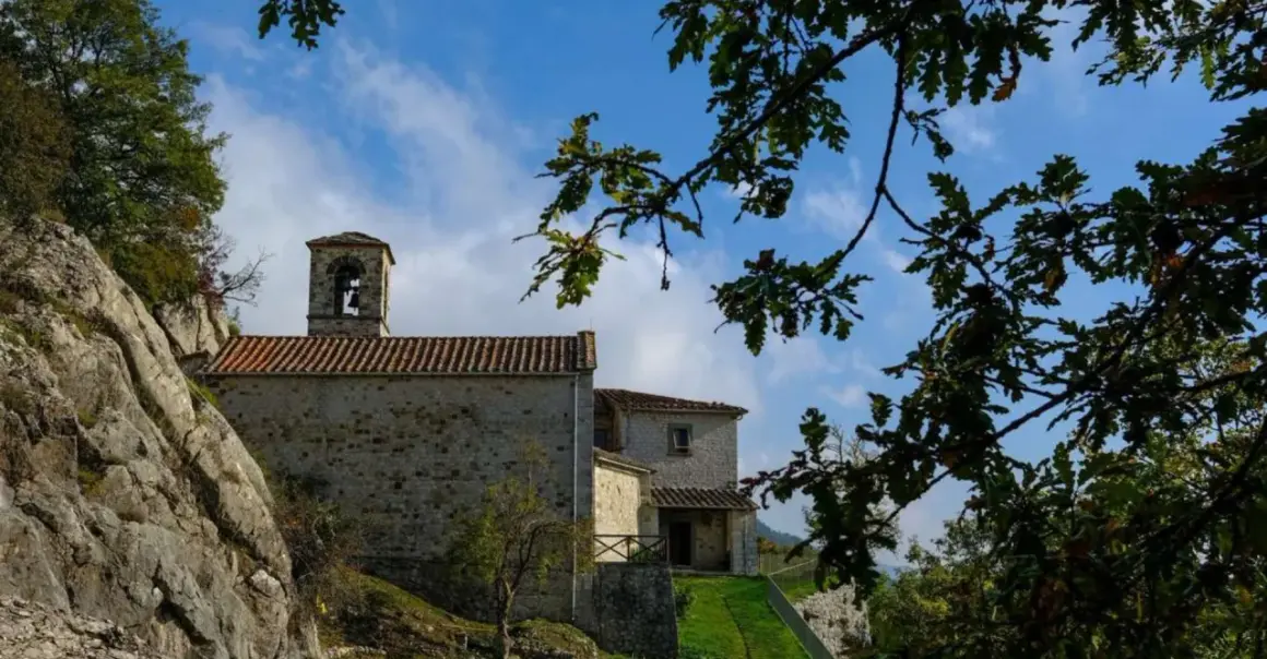 L'eremo di Cerbaiolo, con la sua torre campanaria in pietra, si staglia contro un cielo azzurro tra i rami di una quercia.