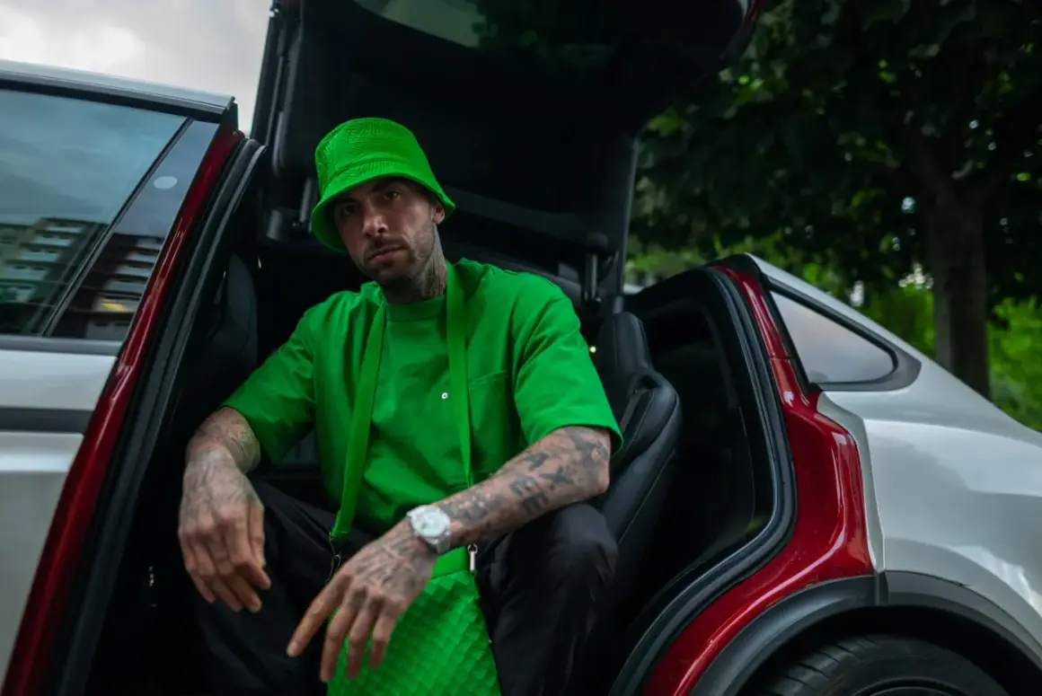 Malik Montana w zielonym kapeluszu i koszulce, siedzi w samochodzie z otwartymi drzwiami. Wygląda jak adwokat diabła, gotowy na nowe wyzwania.