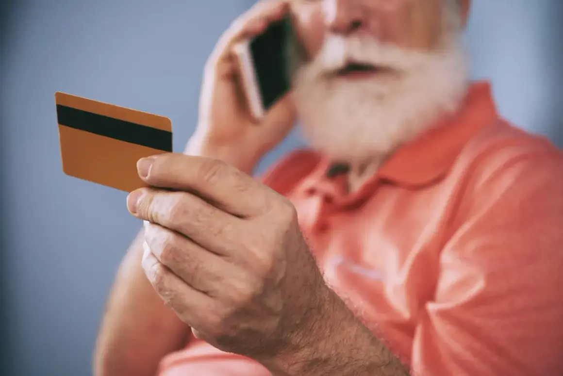 Starszy mężczyzna z brodą trzyma kartę i rozmawia przez telefon, zastanawiając się, dlaczego bank zablokował kartę.