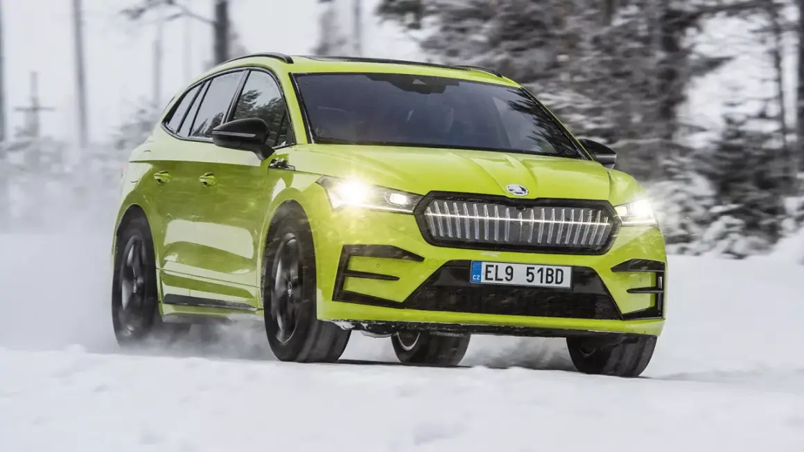 En ljusgrön Skoda Enyaq testas i snö. Bilen kör genom ett vinterlandskap med träd i bakgrunden.