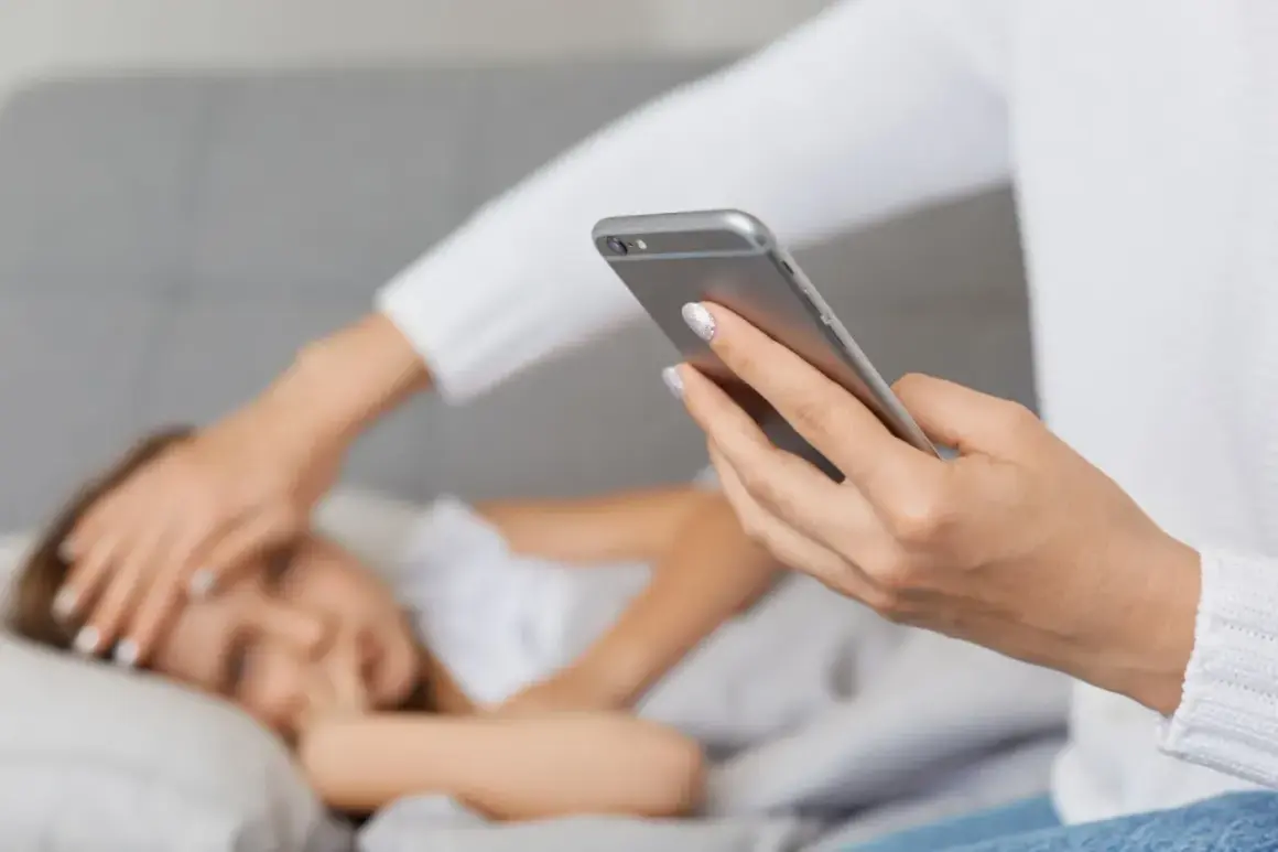 Kobieta z telefonem w dłoni sprawdza gorączkę dziecku leżącemu w łóżku.