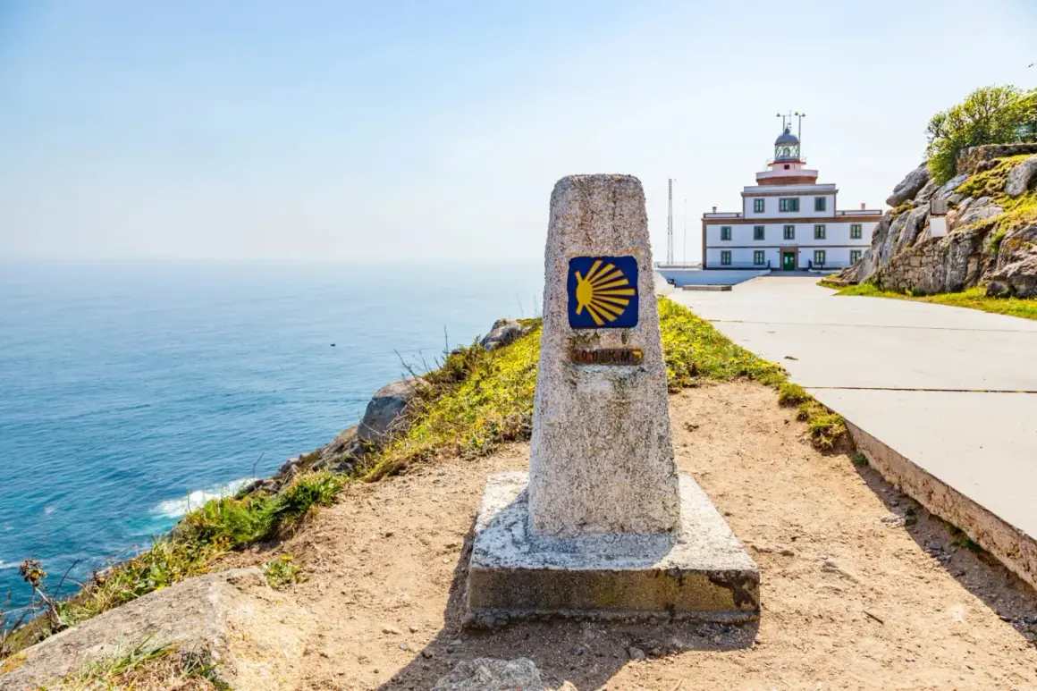 Kamienna wskazówka z symbolem muszli i napisem "0.000 KM" na trasie Camino Portugues. W tle biała latarnia morska i błękitne morze.