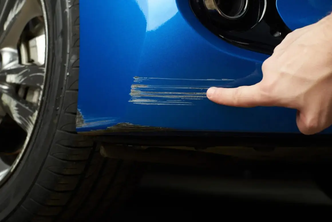 Dito si vede come riparare graffi auto. Un dito indica profondi graffi su un'auto blu vicino alla ruota.