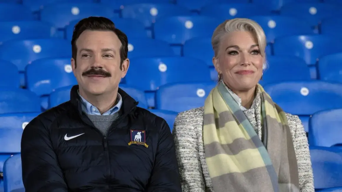 Die Besetzung von Ted Lasso, Jason Sudeikis und Hannah Waddingham, sitzt lächelnd auf blauen Tribünen.