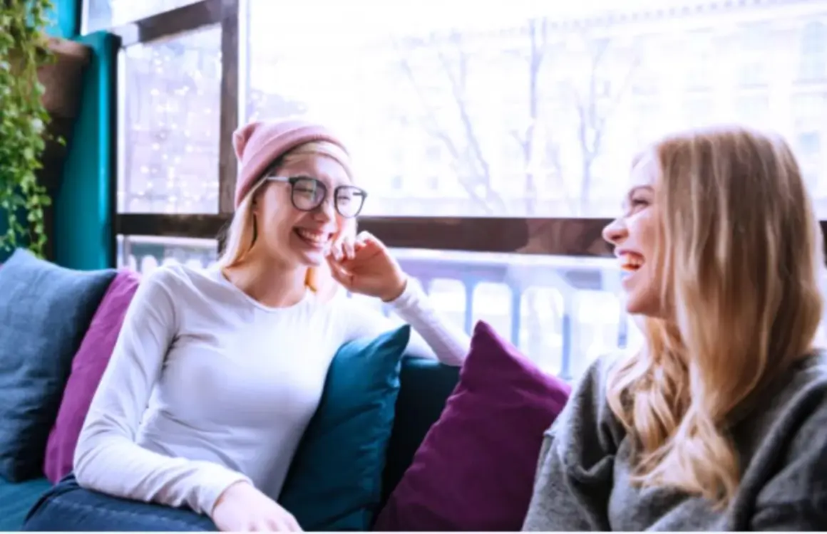 Dos amigas ríen sentadas en un sofá, como si compartieran anécdotas de couchsurfing que es una forma de viajar.