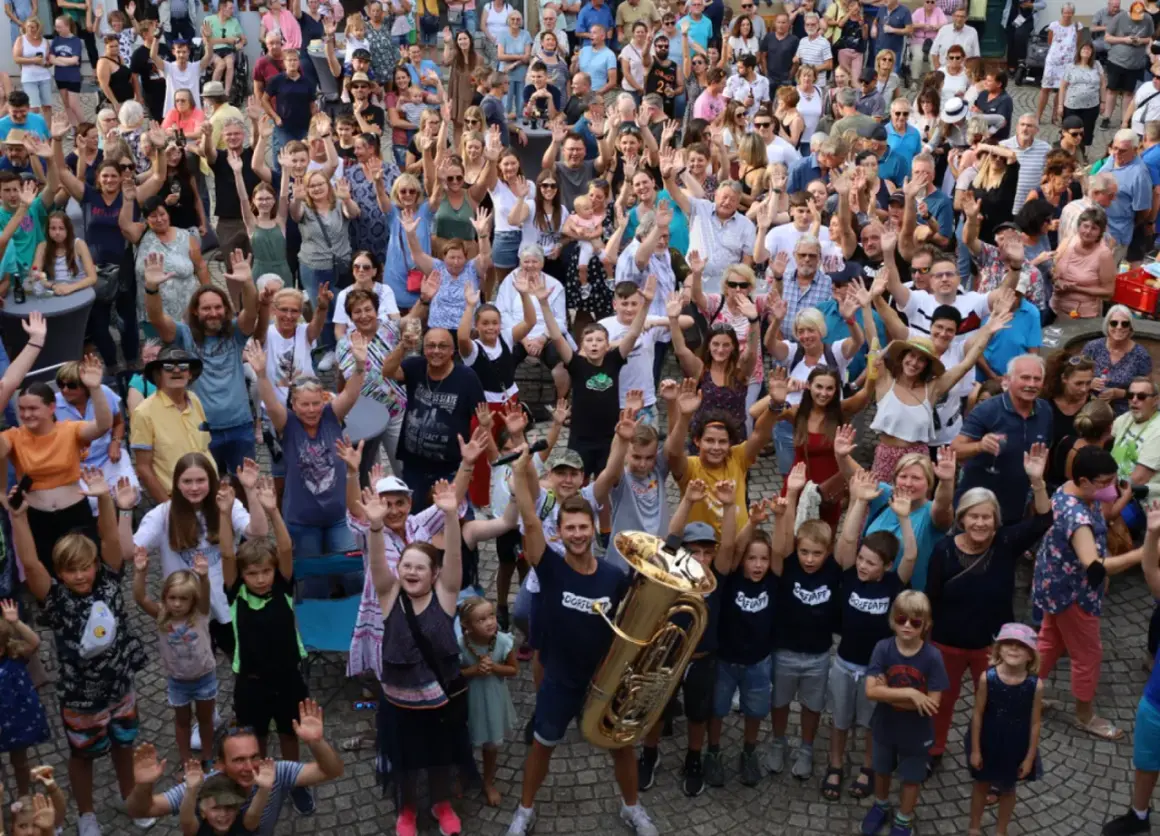 Große Menschenmenge winkt fröhlich bei Brackenheim Veranstaltungen. Ein Mann hält eine Tuba, Kinder und Erwachsene sind begeistert.