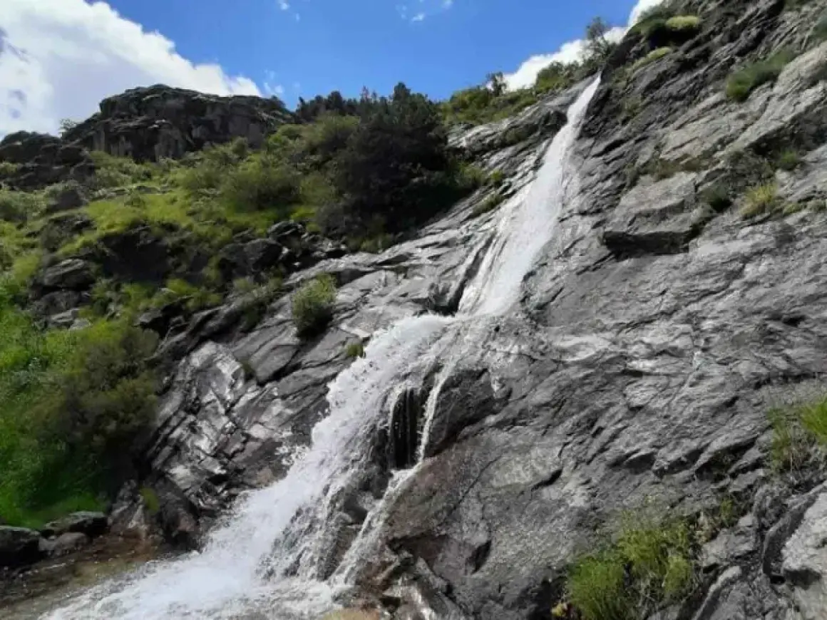 Una hermosa cascada en Madrid, cayendo por rocas grises y vegetación verde bajo un cielo azul con nubes blancas.