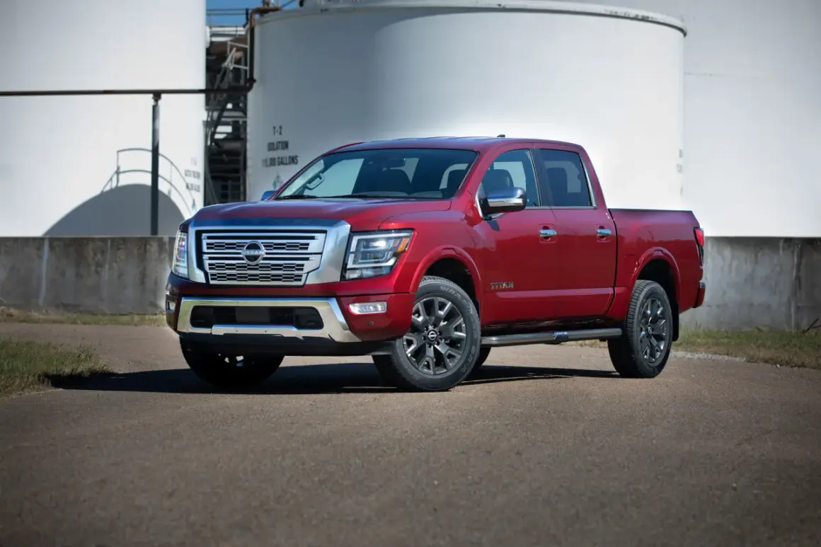 Czerwony Nissan Titan z chromowanymi detalami stoi na asfalcie przed białymi zbiornikami przemysłowymi.