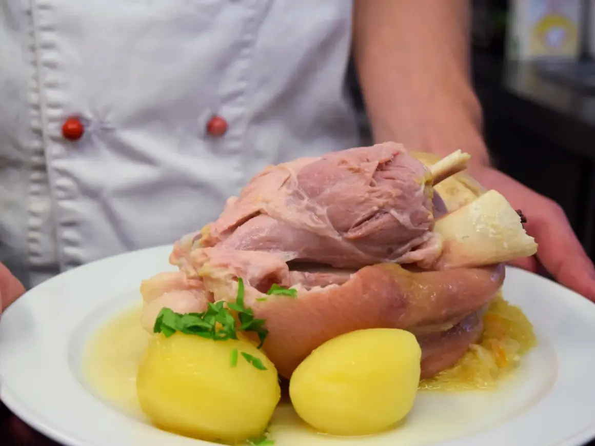 Ein Koch präsentiert ein deftiges Eisbein mit Kartoffeln und Sauerkraut. Die Frage "Eisbein wie lange kochen?" beschäftigt viele Hobbyköche.