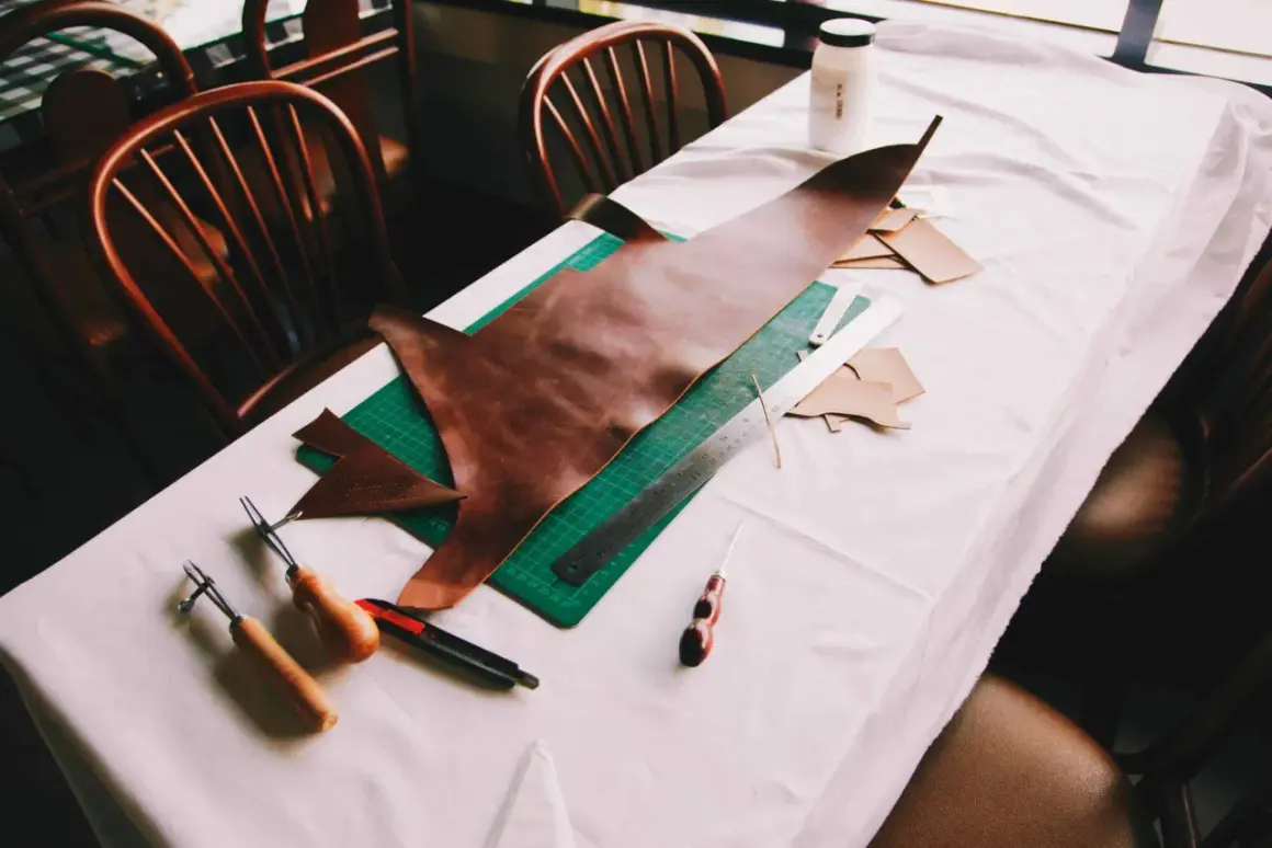 Sur une table recouverte d'une toile blanche, des outils de maroquinerie et des pièces de cuir sont disposés. On y voit un grand morceau de cuir brun, une planche à découper verte, une règle, un cutter, et des outils pour travailler le cuir, suggérant ...