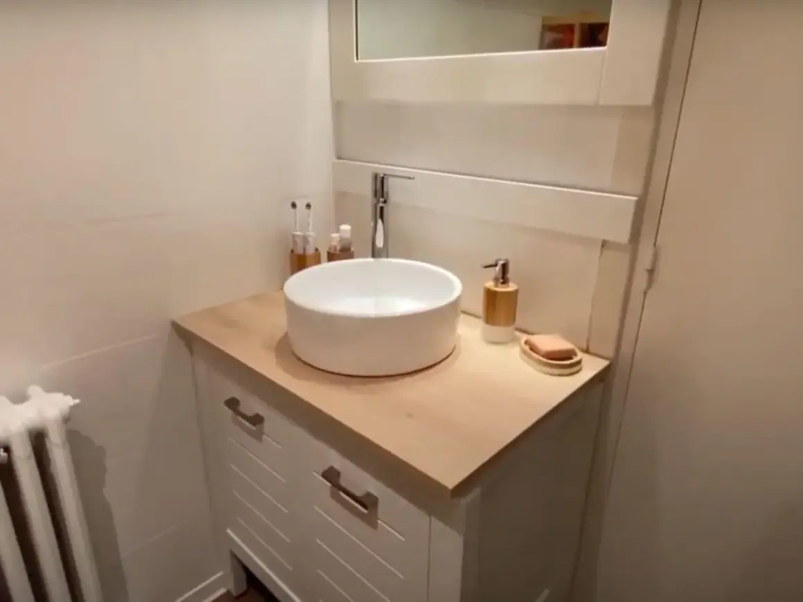 Un meuble de salle de bain relooké avec un plan de travail en bois clair, une vasque blanche et des accessoires modernes.