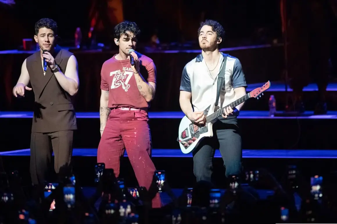 Trzech członków zespołu Jonas Brothers na scenie, jeden śpiewa, drugi gra na gitarze.