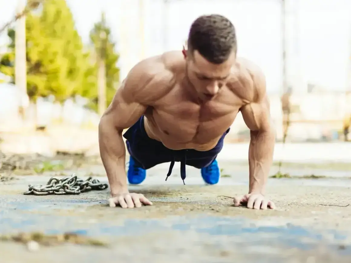 Mięśniowy mężczyzna wykonuje pompki na biceps na zewnątrz. Obok leży ciężki łańcuch.
