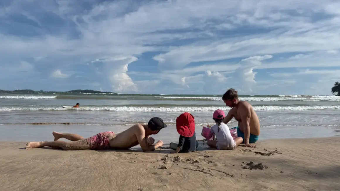 Familj njuter av en solig dag på stranden i Sri Lanka. En man ligger i sanden medan en annan leker med barnen vid havet.
