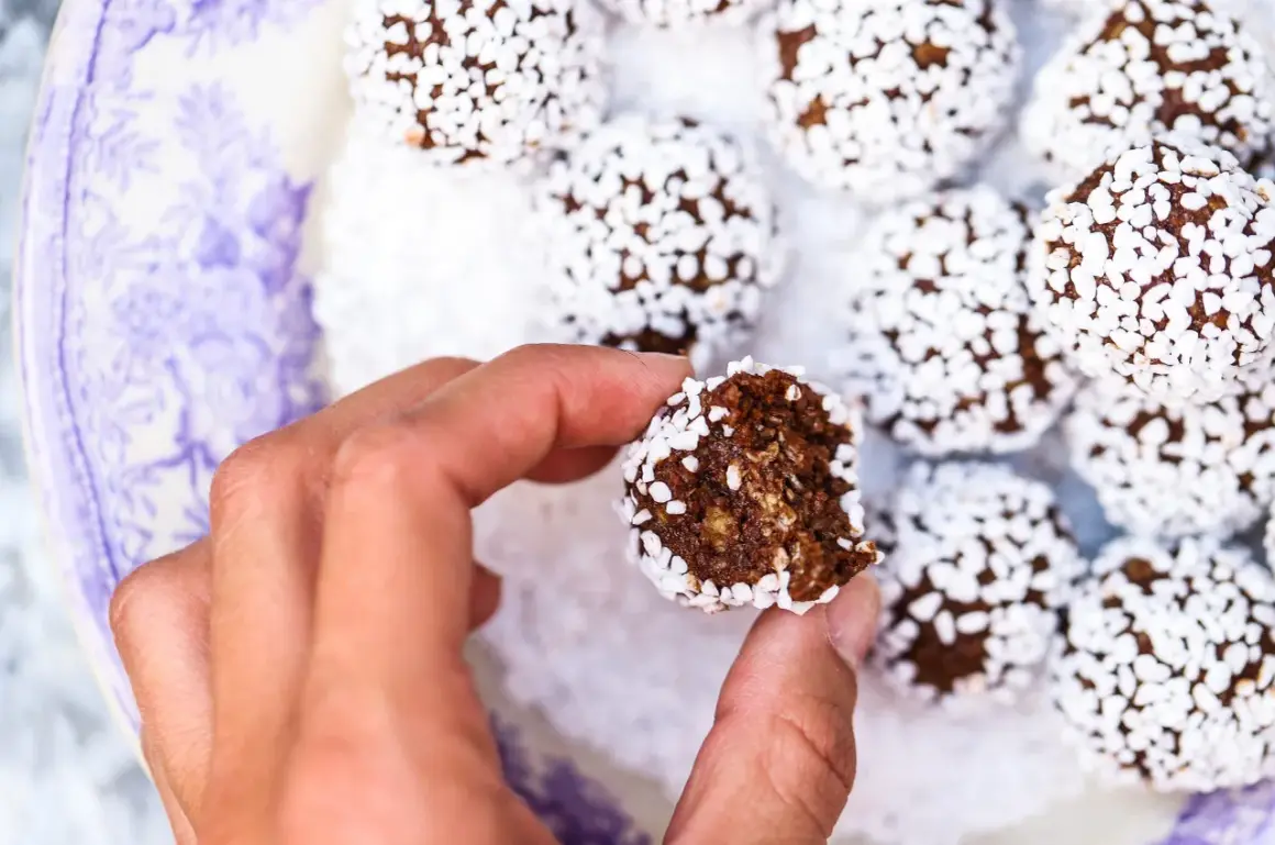 En hand håller en bit av nyttiga chokladbollar, rullade i kokosflingor, på en vacker lila-vit tallrik.