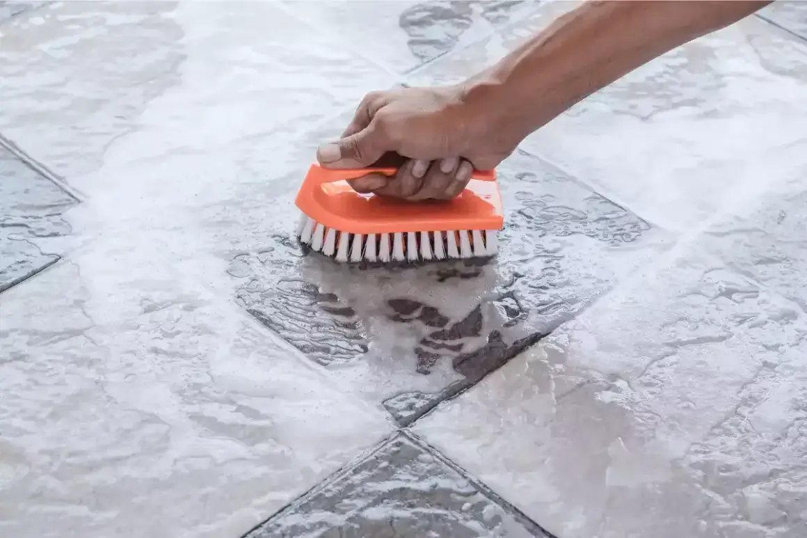 Main tenant nettoie une tache acide sur carrelage avec une brosse orange.