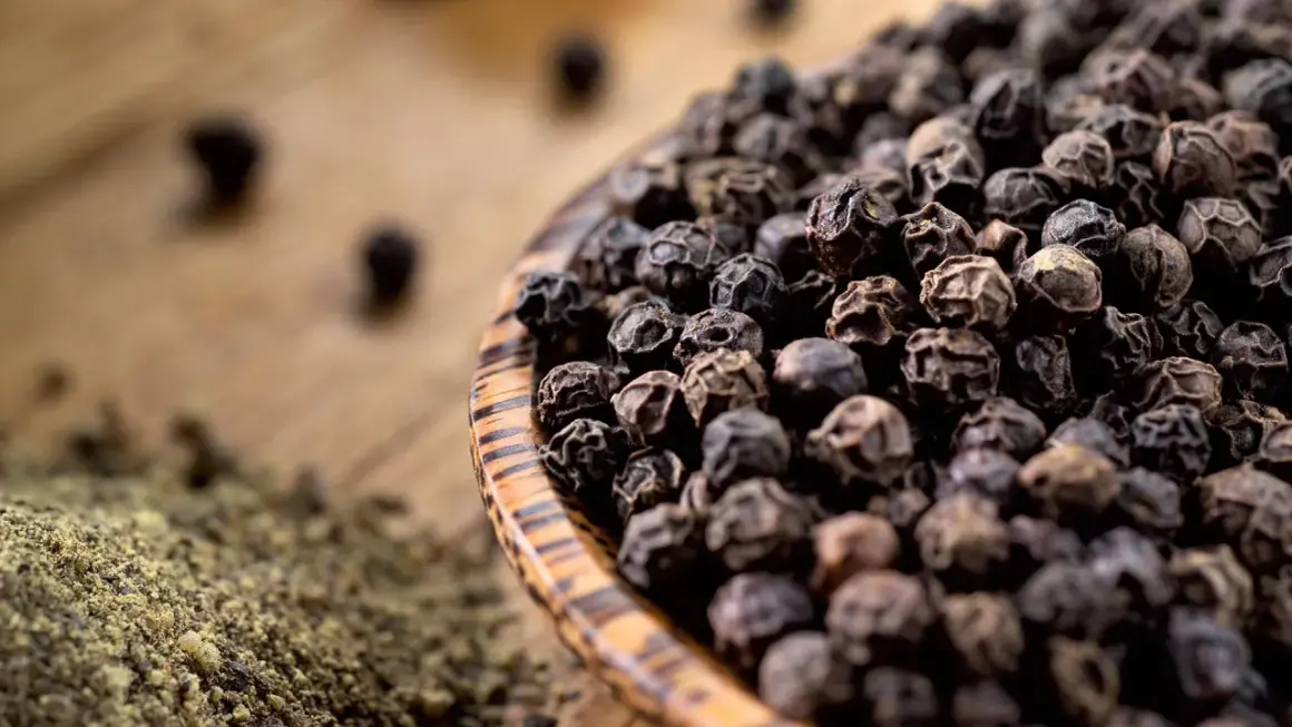 Schwarze Pfefferkörner in einer Holzschale, daneben gemahlener Pfeffer. So wird Pfeffer hergestellt: Ernte, Trocknung und Mahlung.