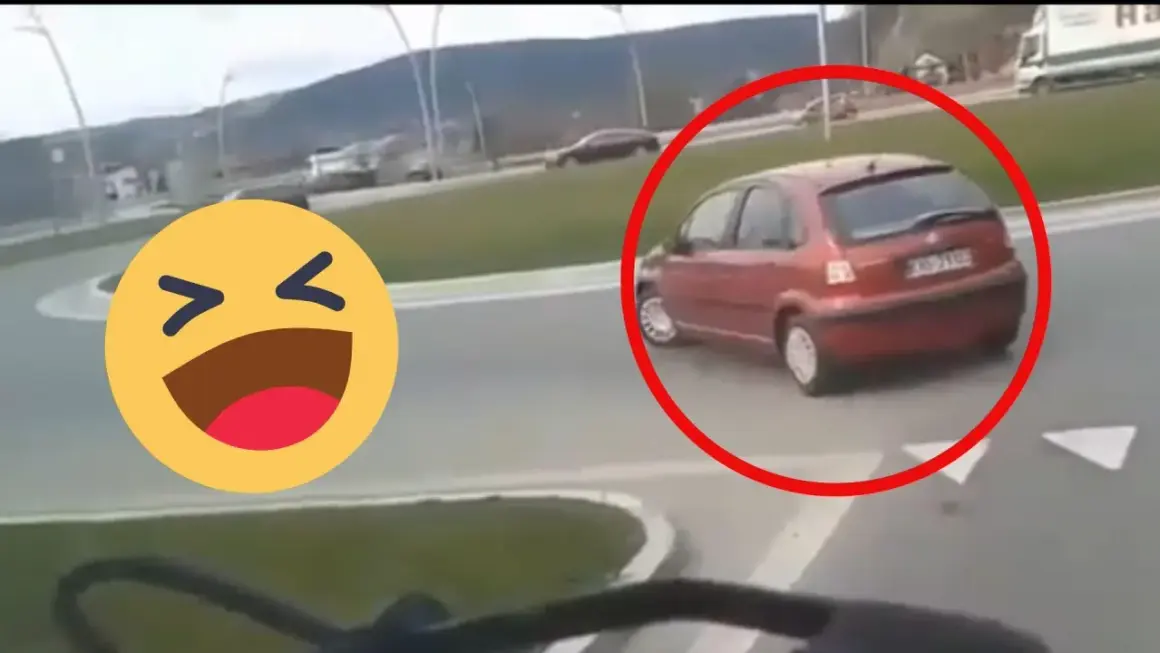 Czerwony samochód próbuje zawrócić na rondzie, ale robi to na lewo, co wygląda na niezbyt przemyślane. Obok śmieje się emotikon.