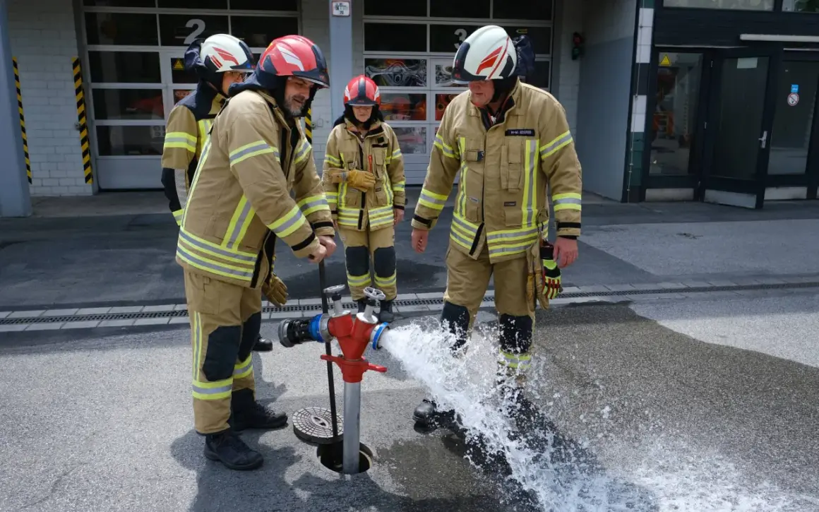 Jugendfeuerwehr übt den Umgang mit dem Hydranten. Wasser spritzt aus dem roten Standrohr, während die jungen Brandschützer in voller Montur zuschauen.