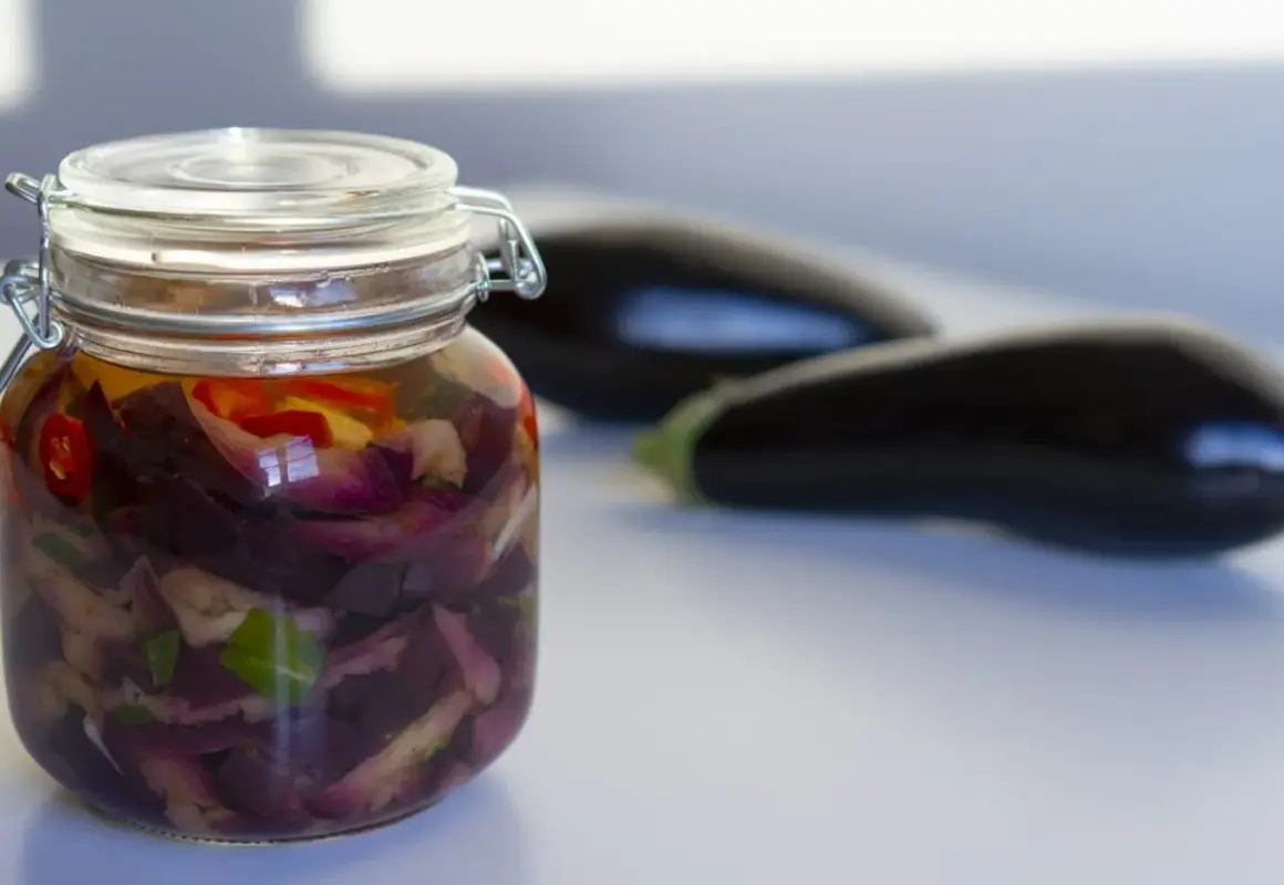Berenjenas en escabeche, cortadas y marinadas en un frasco de vidrio, con berenjenas frescas desenfocadas al fondo.