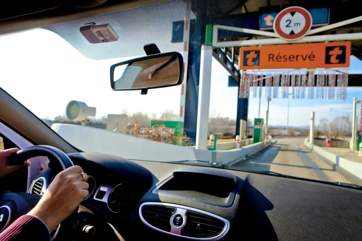 Vue depuis l'intérieur d'une voiture approchant une voie de péage. Le tableau de bord et le volant sont visibles. Un badge de télépéage est fixé au pare-brise. Le panneau "Réservé" indique comment ça fonctionne.