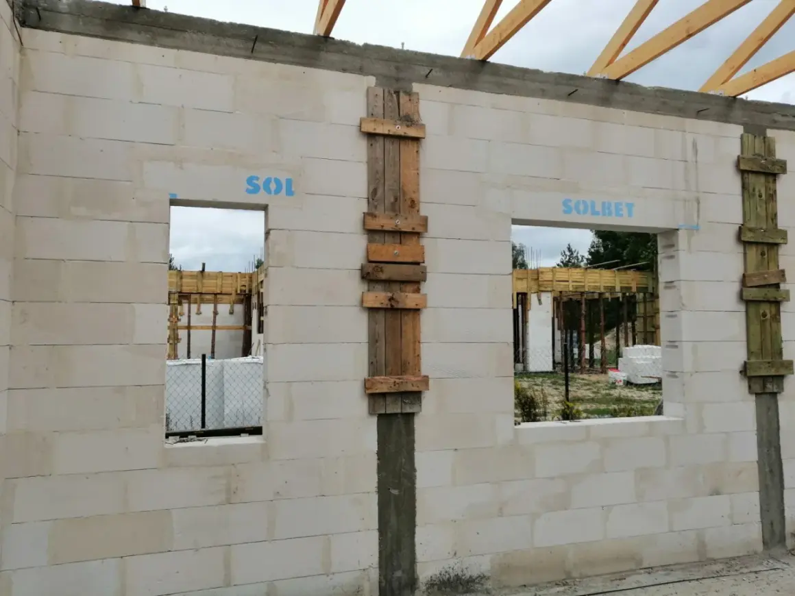 Budowa domu z bloczków betonowych, widoczne okna i drzwi z tymczasowym nadprożem, drewniane elementy konstrukcyjne.