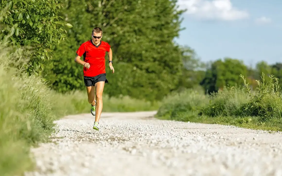 Mann in rotem Shirt und schwarzen Shorts absolviert einen Fartlek Lauf auf einem Schotterweg, umgeben von grüner Natur.
