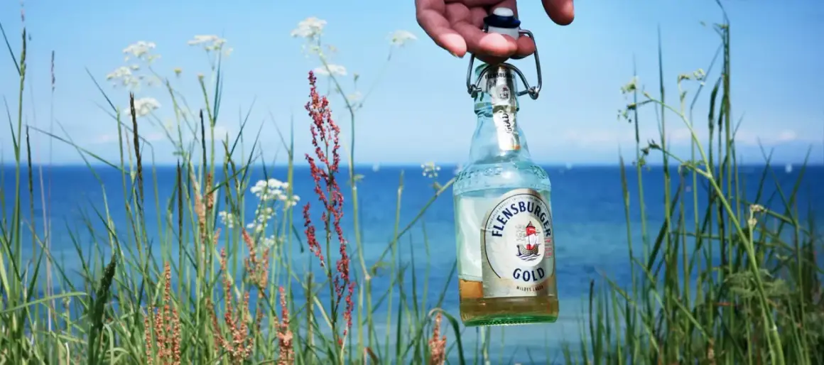 Eine Flensburger Gold Flasche wird vor der Küste gehalten. Der leichte Bier Geschmack passt perfekt zum Sommer.