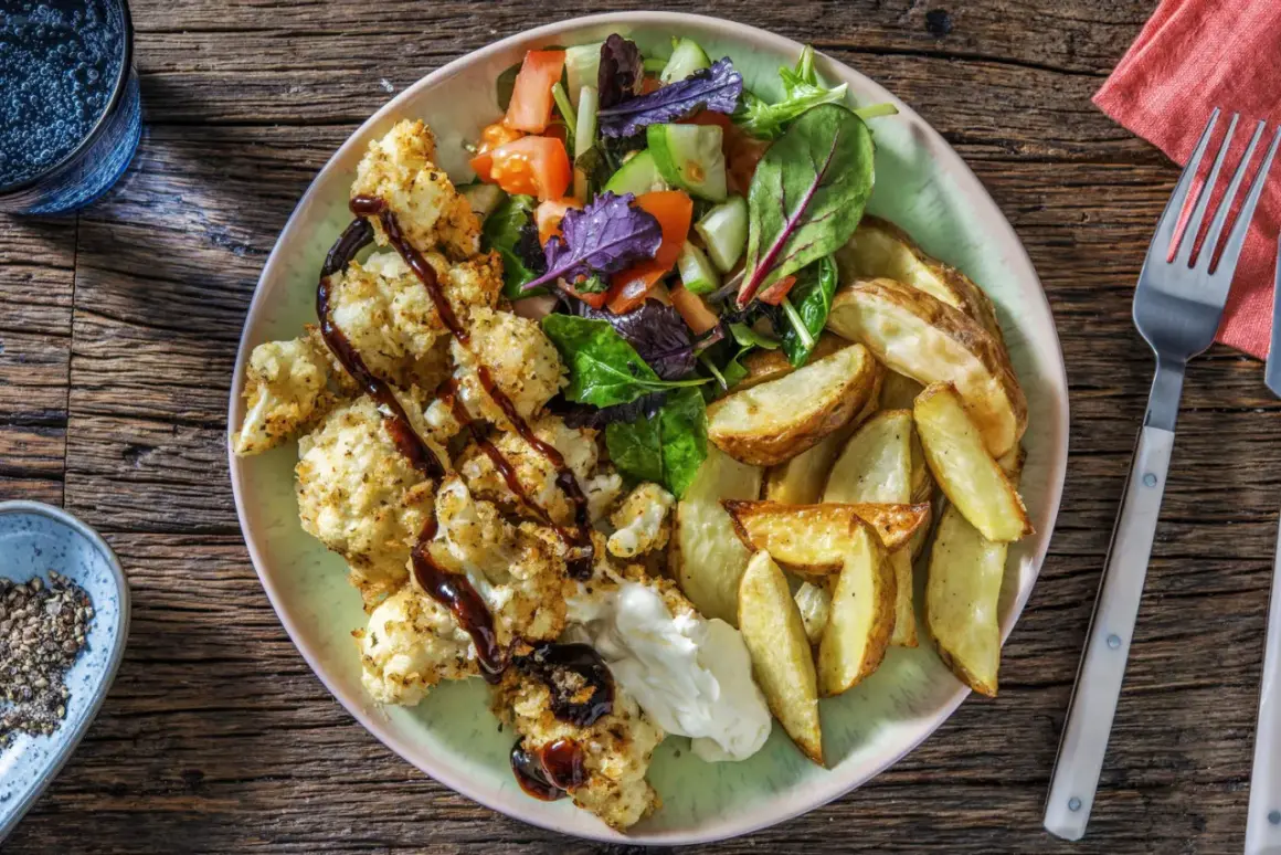 En tallrik med rostad blomkål i ugn, klyftpotatis och en fräsch sallad. Blomkålen är täckt med ströbröd och ringlad med en mörk sås.