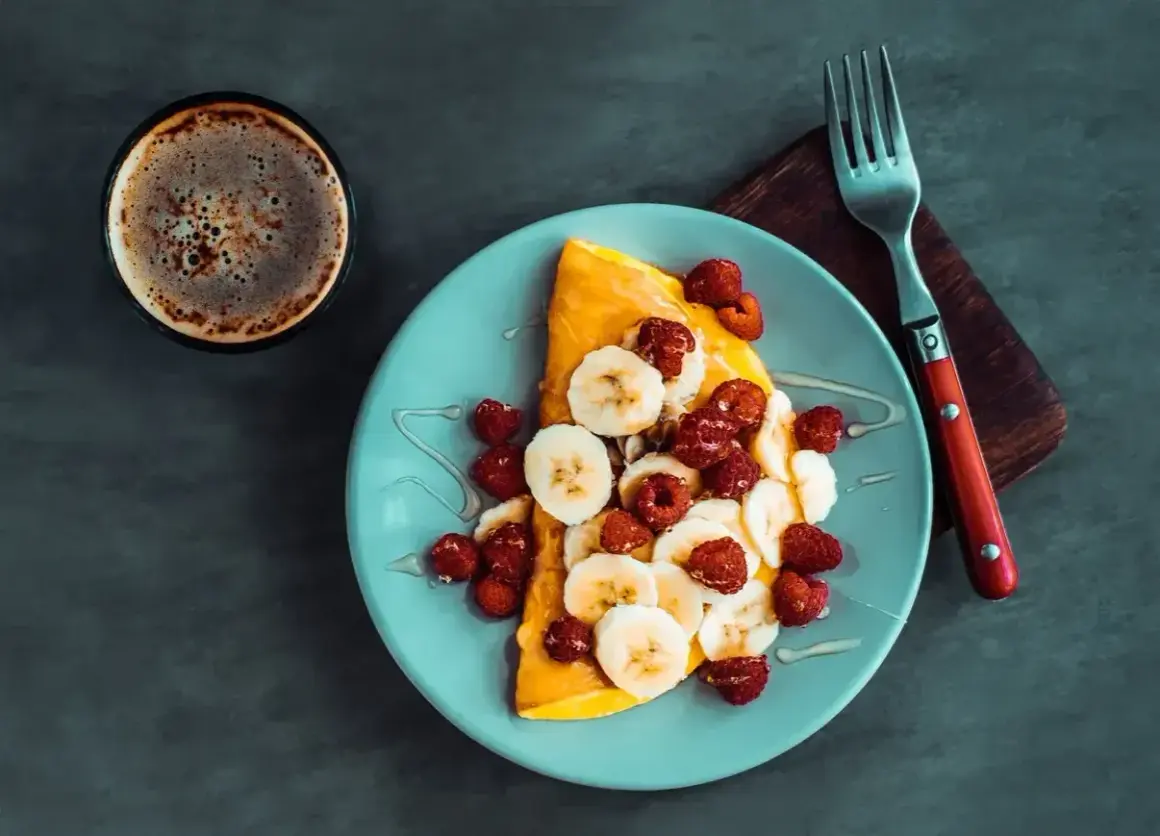Słodki omlet z bananami i malinami, polany syropem, obok filiżanka kawy. Idealny na słodki początek dnia.