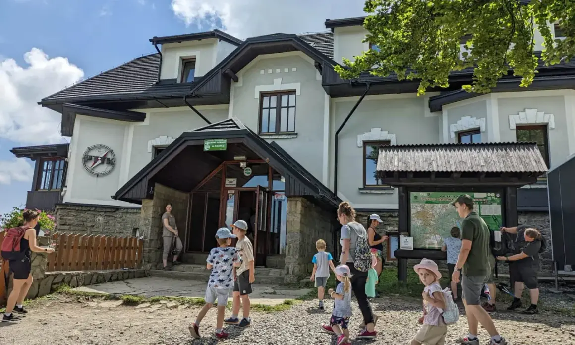 Rodzina przy schronisku na Magurce Wilkowickiej. Dzieci zwiedzają okolicę, a dorośli planują trasę na mapie.