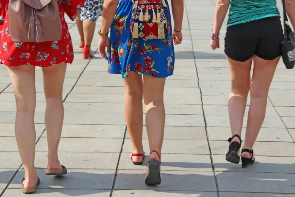 Trzy kobiety idą chodnikiem. Jedna ma na sobie czerwoną sukienkę w kwiaty, druga niebieską, a trzecia czarne szorty. Widać zaczerwienione obtarte uda u kobiety w czerwonej sukience.