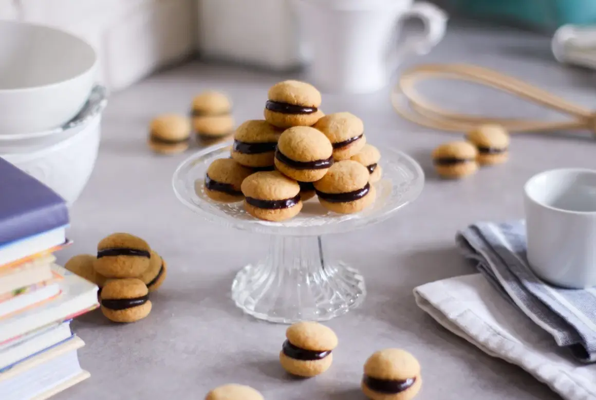 Baci di dama, deliziosi biscotti con crema al cioccolato, disposti su un'alzata di vetro, circondati da libri e tazze.