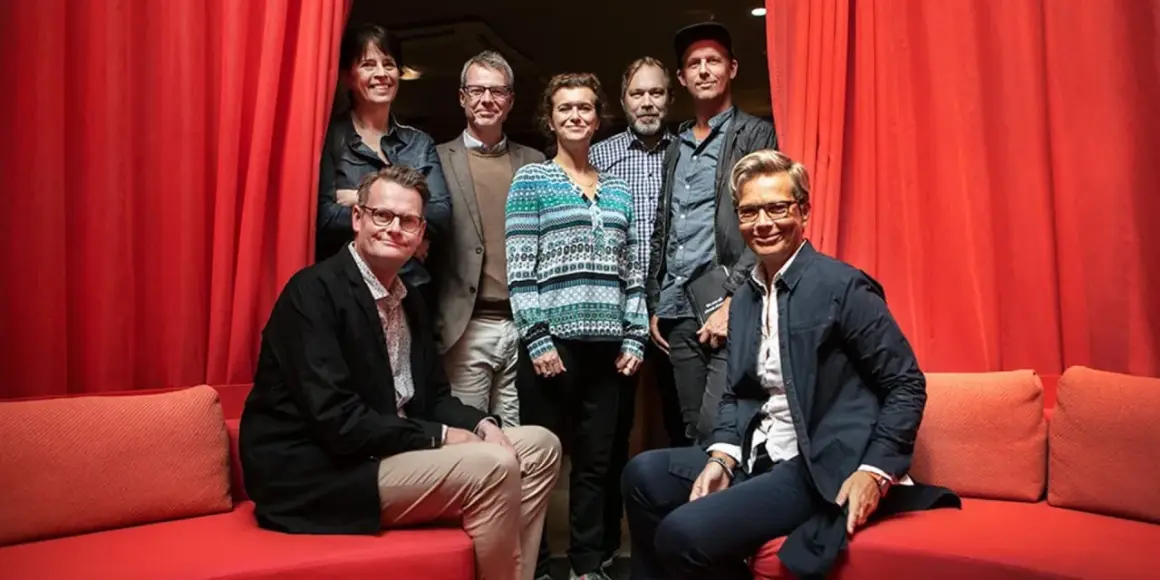 Gruppbild med åtta personer framför röda draperier. Izabella Jensen står i mitten, klädd i en mönstrad tröja.