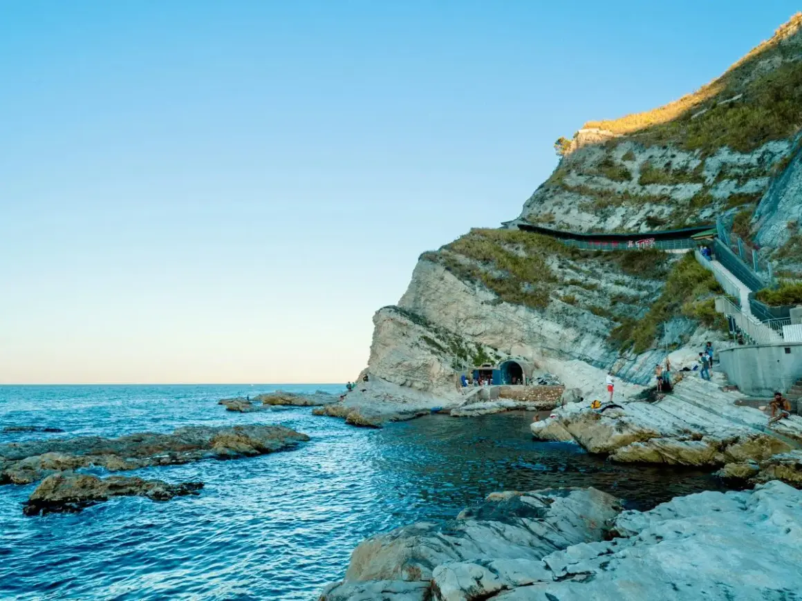 Scogliere rocciose e mare blu intenso al Conero, con un tunnel scavato nella roccia e persone che si godono la giornata.