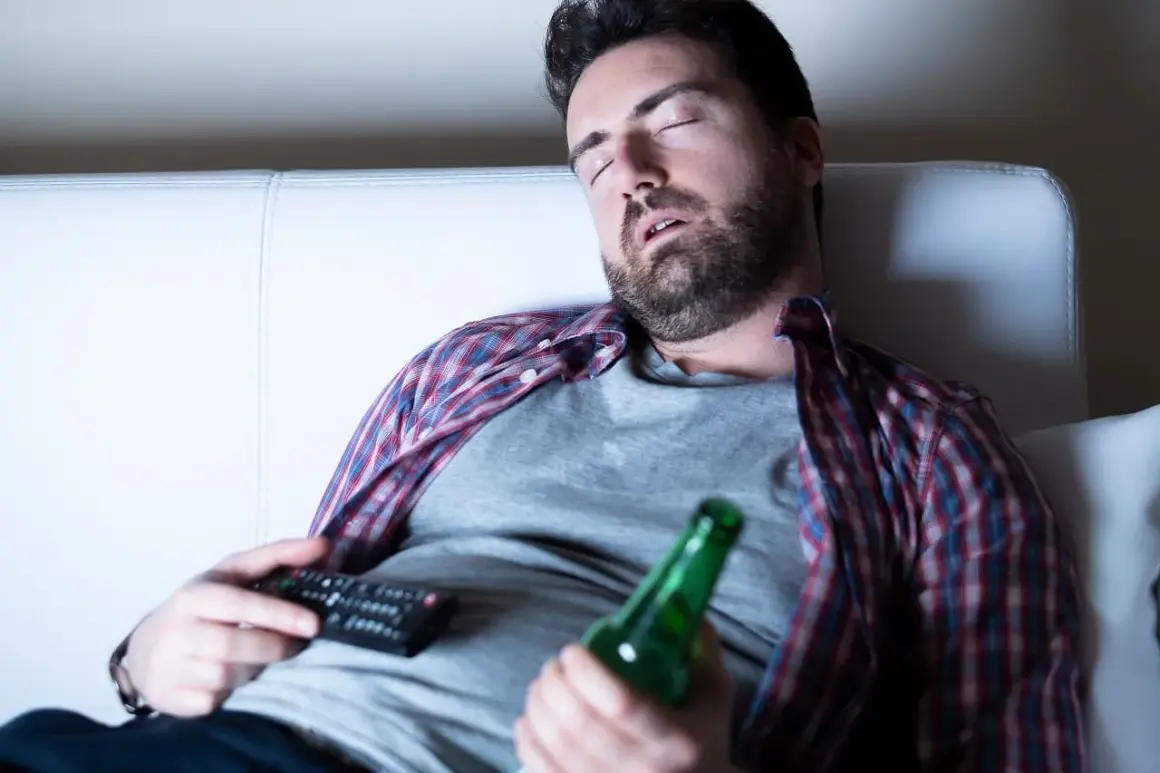 Mężczyzna śpi na kanapie z pilotem i butelką piwa, być może doświadczając dziwnych snów po odstawieniu alkoholu.