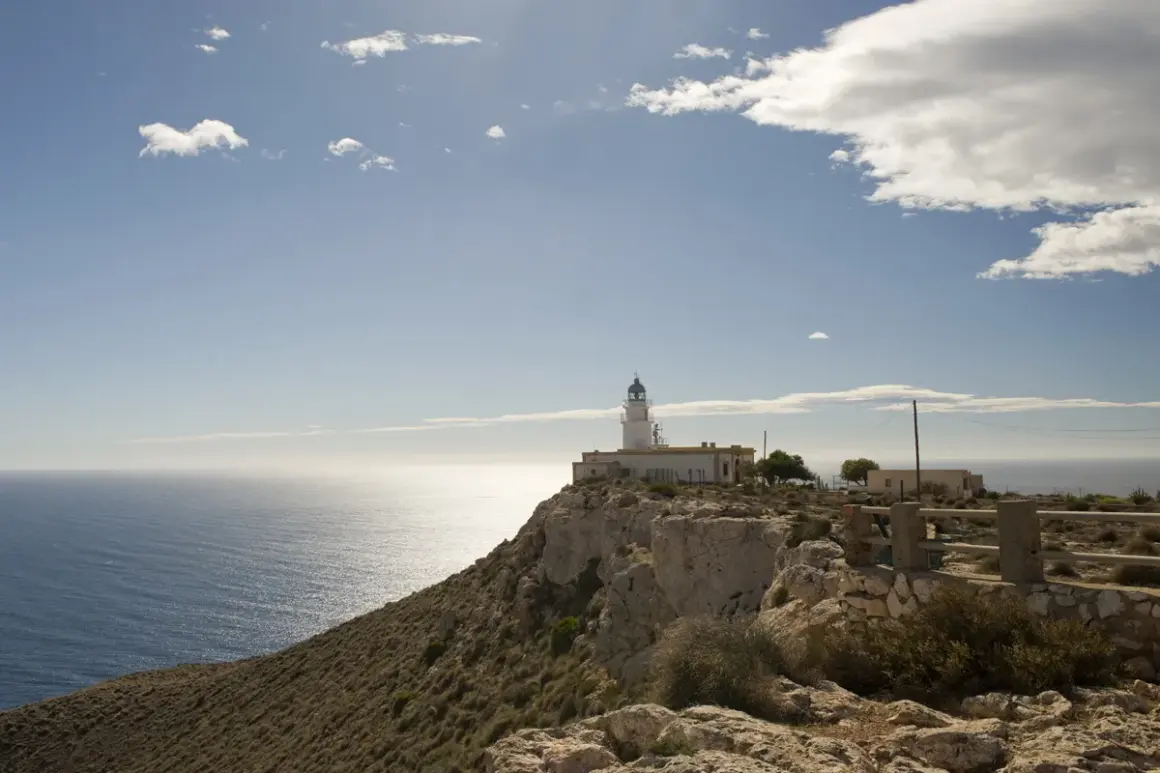El faro de Mesa Roldán se alza sobre un acantilado rocoso, vigilando el mar Mediterráneo bajo un cielo azul con nubes dispersas.