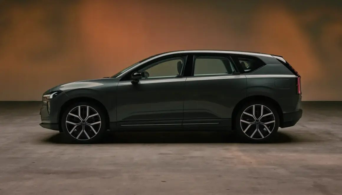 Volvo EX60 gris oscuro, perfil lateral, con fondo degradado naranja y suelo de hormigón.
