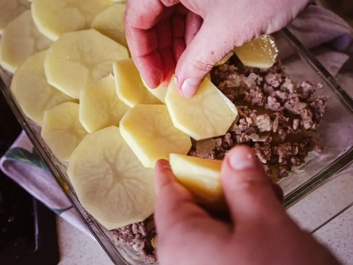 Ręce układają plastry ziemniaków na warstwie mięsa mielonego, tworząc pyszną zapiekankę ziemniaczaną z mięsem mielonym.