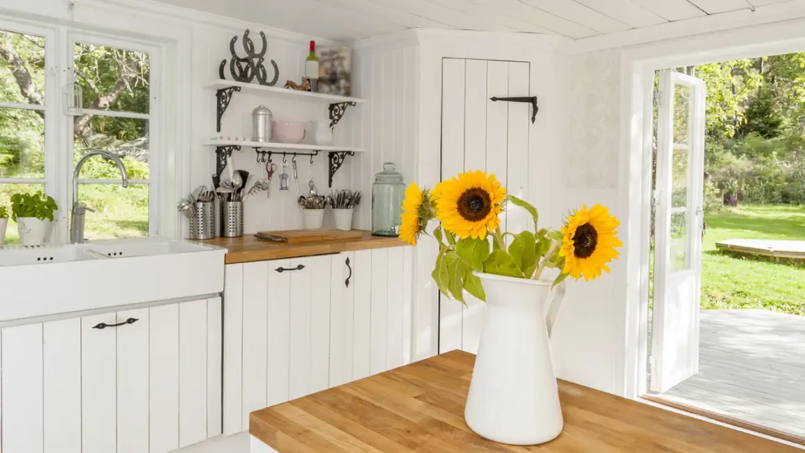 Helle Küche im modernen Landhausstil mit Spülbecken, Holzarbeitsplatte und Sonnenblumen. Offene Tür gibt Blick auf grünen Garten frei.