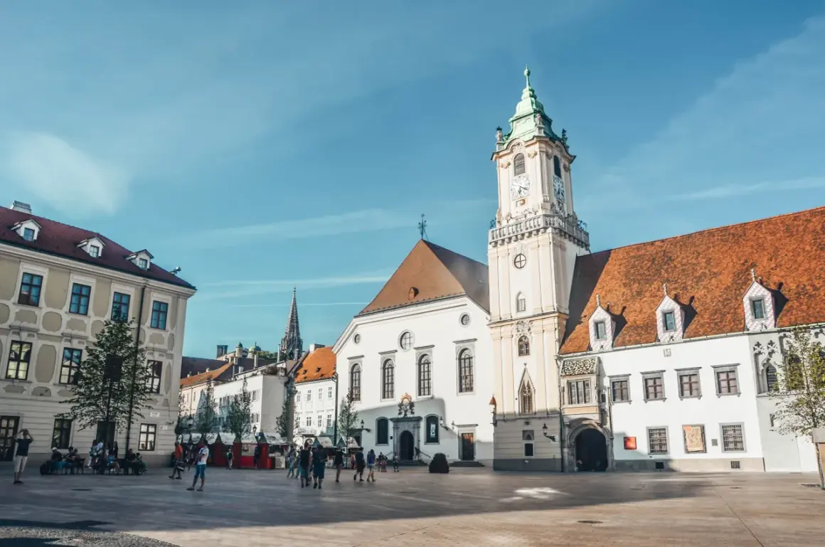 Słoneczny dzień na bratysławskim rynku. Ludzie spacerują po brukowanym placu przed zabytkowymi budynkami z charakterystyczną wieżą zegarową.