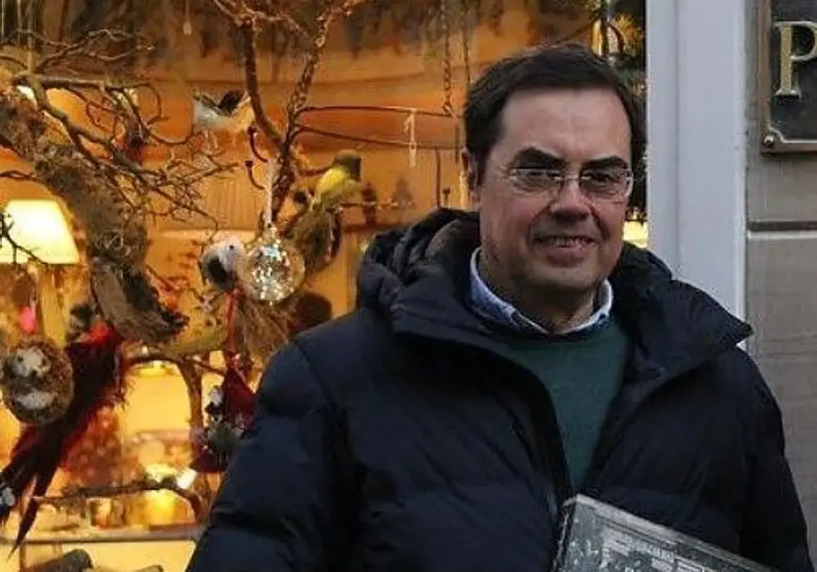 Cayo Pombo Ybarra, sonriendo, con un abrigo oscuro y un suéter verde, frente a un escaparate decorado con pájaros y adornos.