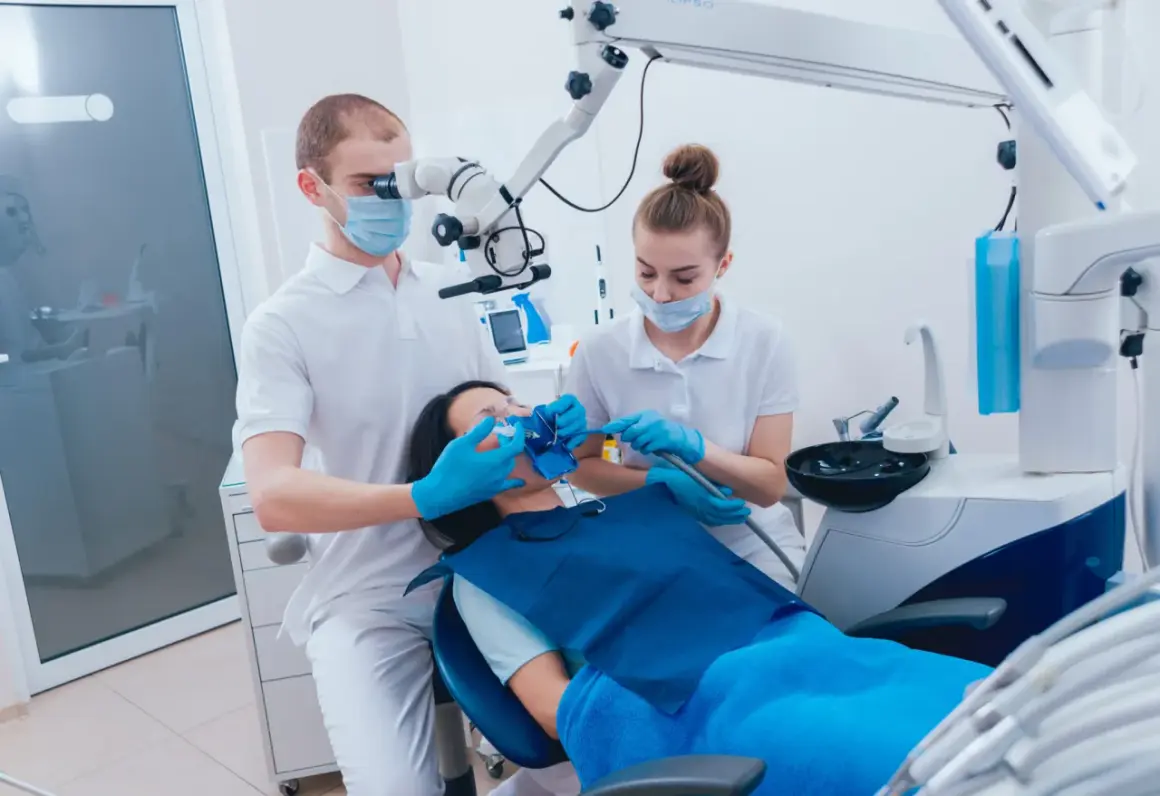 Dentysta pod mikroskopem wykonuje ekstyrpację miazgi u pacjentki. Asystentka pomaga.