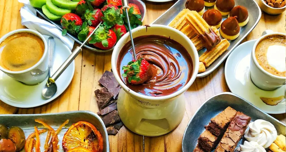 Jak przygotować czekolady do fontanny: truskawka zanurzona w płynnej czekoladzie, obok kawa, ciastka, owoce i brownie.