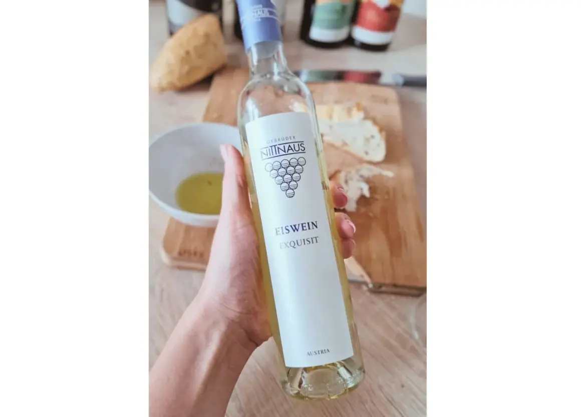 Dłoń trzyma butelkę lodowego wina Eiswein Exquisit z Austrii. Na stole leży chleb i miseczka z oliwą.