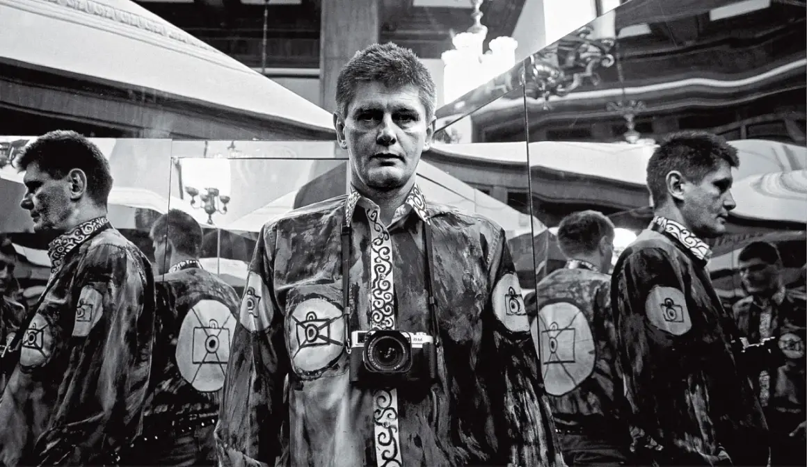Krzysztof Miller w lustrzanym odbiciu, z aparatem na szyi, w koszuli z motywem oka.