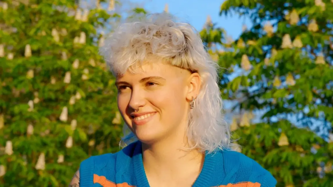 Ragazza con un audace mullet donna lungo, capelli biondi cotonati e rasati ai lati, sorride con gioia sotto un cielo azzurro, circondata da alberi in fiore.