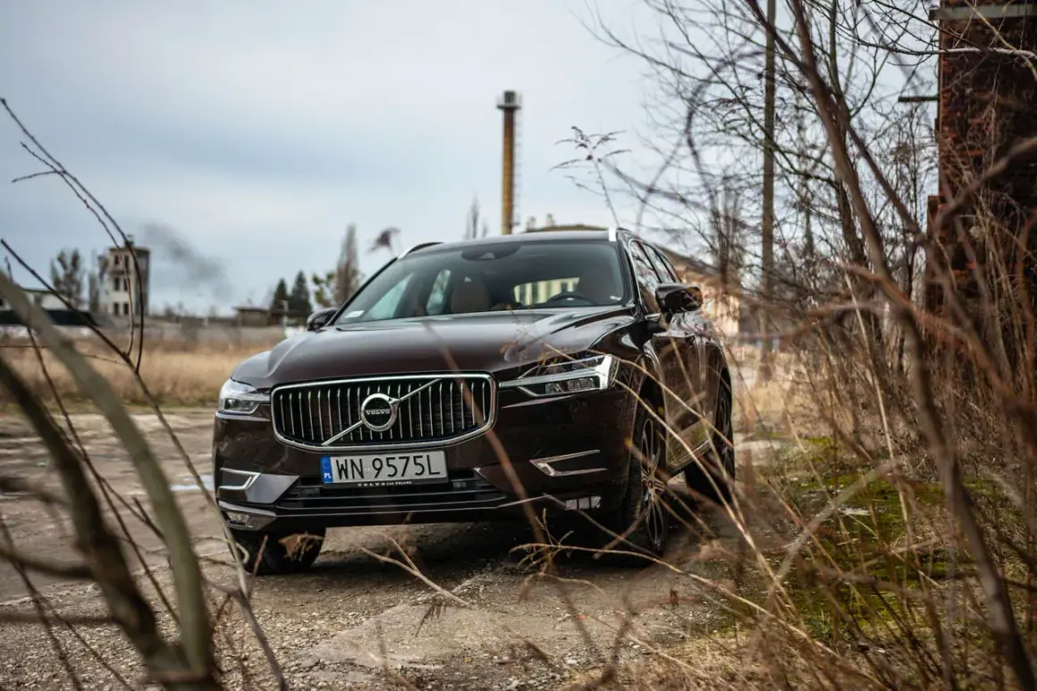 Brązowe Volvo XC60 T5 na tle opuszczonej fabryki i suchych gałęzi.