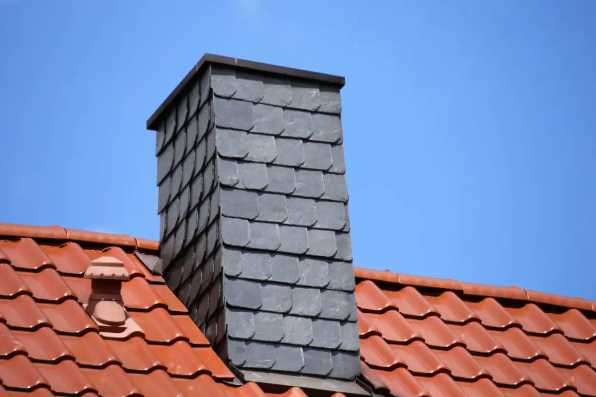 Moderner gemauerter Schornstein verkleidet mit grauen Schieferplatten, überragt ein rotes Ziegeldach unter blauem Himmel.